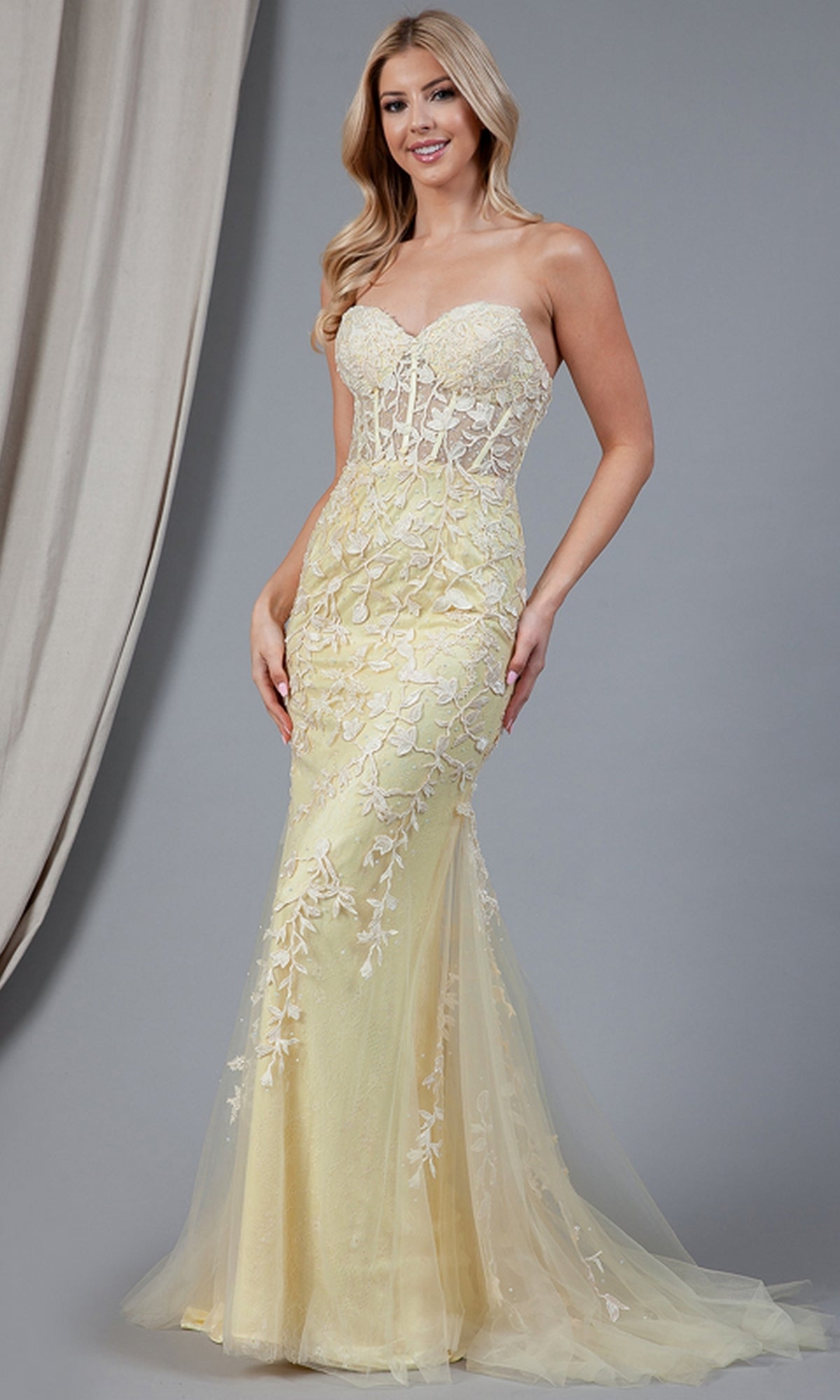 A woman stands against a plain gray background wearing the Strapless Sweetheart Long Mermaid Prom Dress 7024—featuring a fitted, pale yellow corset-style bodice, embroidered florals, and a sheer train.